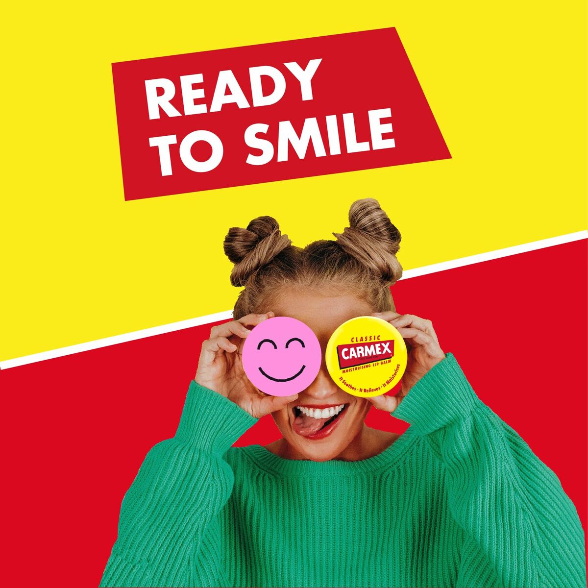 Woman holding Carmex lip balm with a smiley face, wearing a green sweater, against a vibrant yellow and red background.