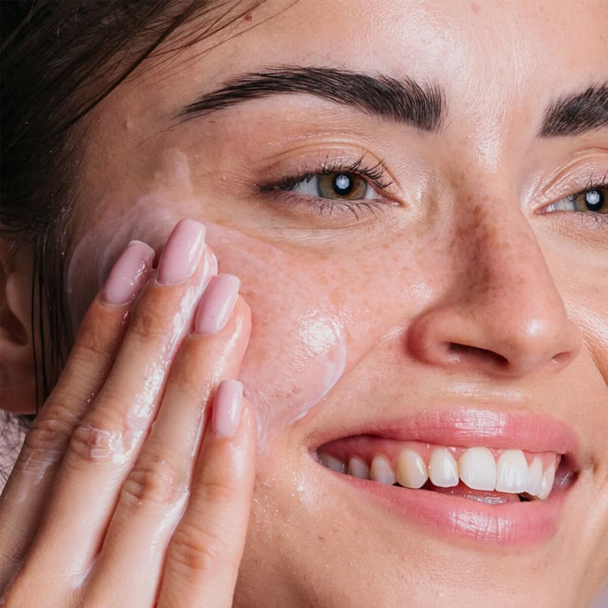 Close-up of a woman smiling while applying a cleanser to her face, showcasing healthy, glowing skin.