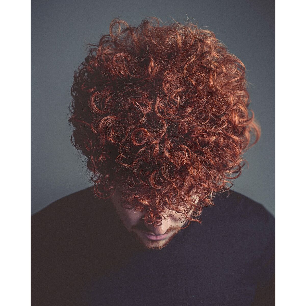 Homme aux cheveux bouclés et roux, regardant vers le bas, avec un fond neutre.
