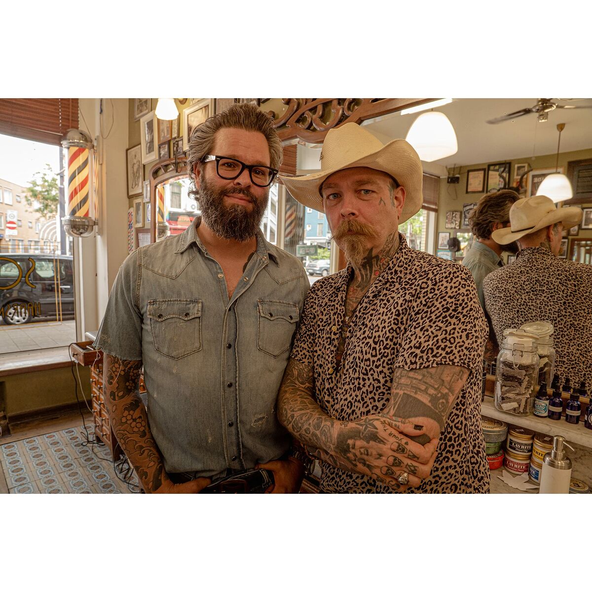 Two tattooed barbers posing in a vintage barber shop with mirrors and hair products.