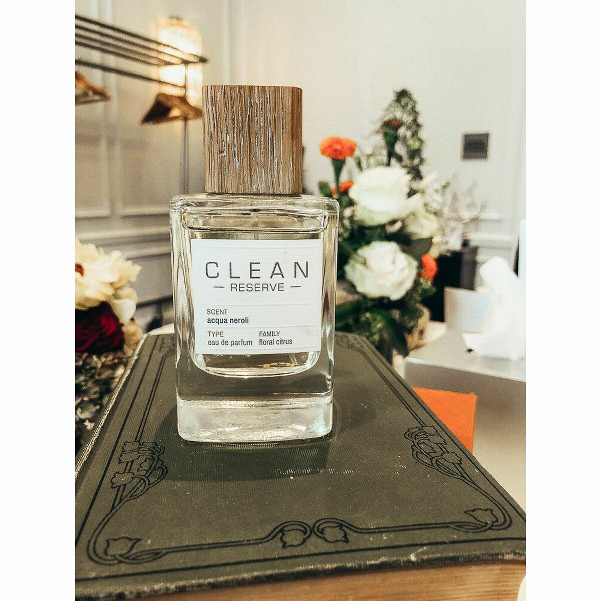 Parfum Unisexe Clean Acqua Neroli EDP 100 ml sur un livre, avec bois sur le bouchon et fleurs en arrière-plan.