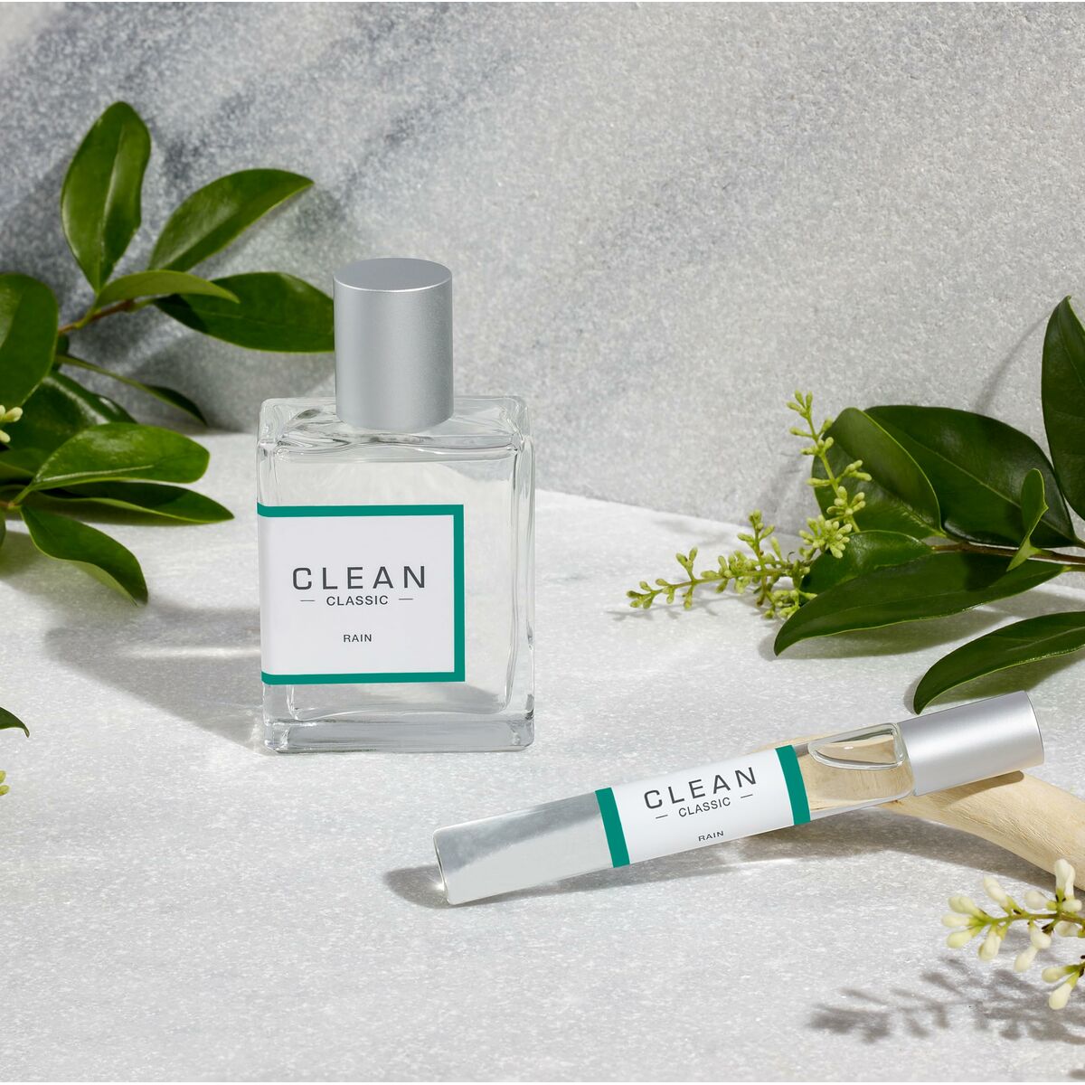 Bottles of CLEAN Rain Eau de Parfum and rollerball on a stone surface surrounded by green leaves.