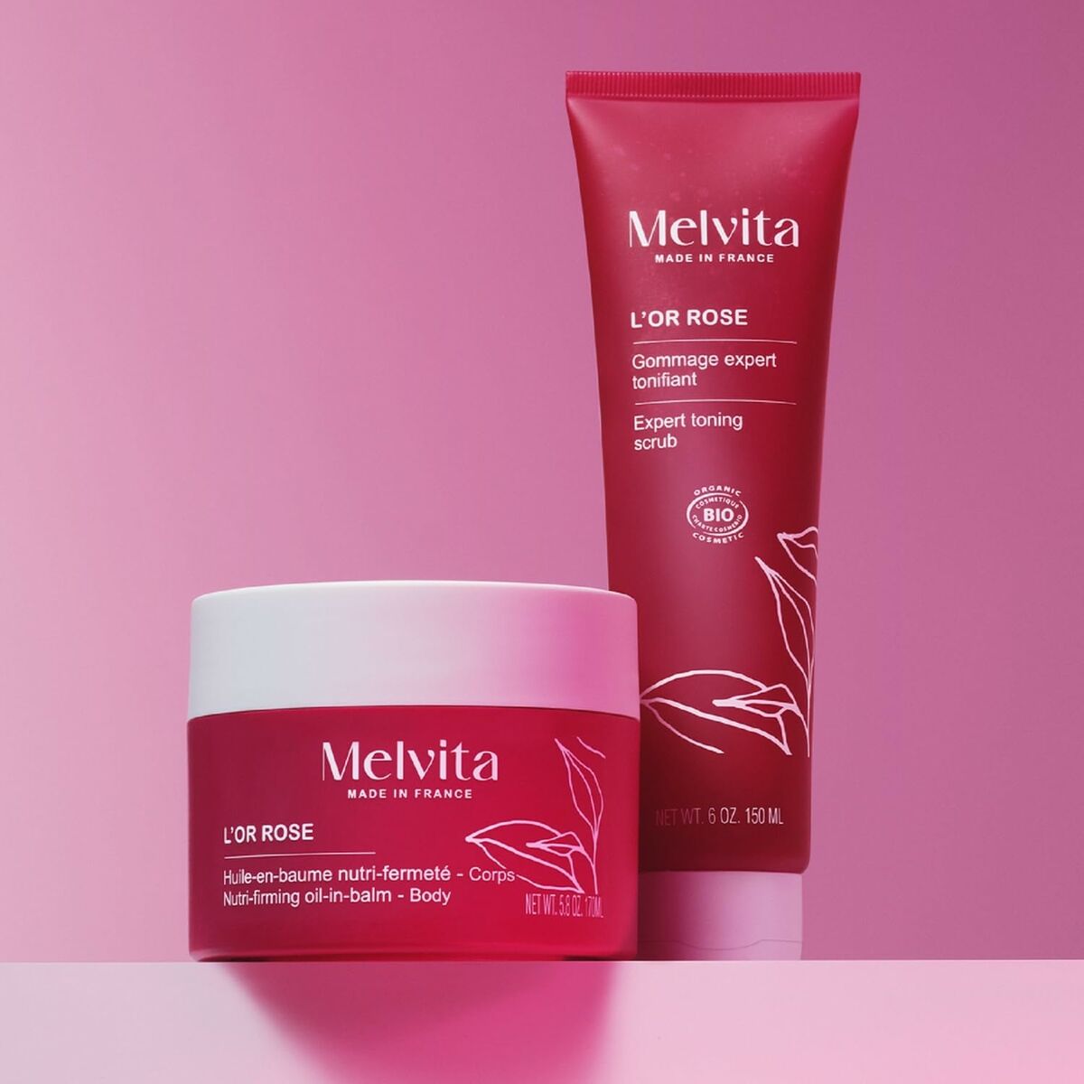 Melvita L'OR ROSE body care products, including a nutri-firm oil-in-balm and an expert toning scrub, on a pink background.