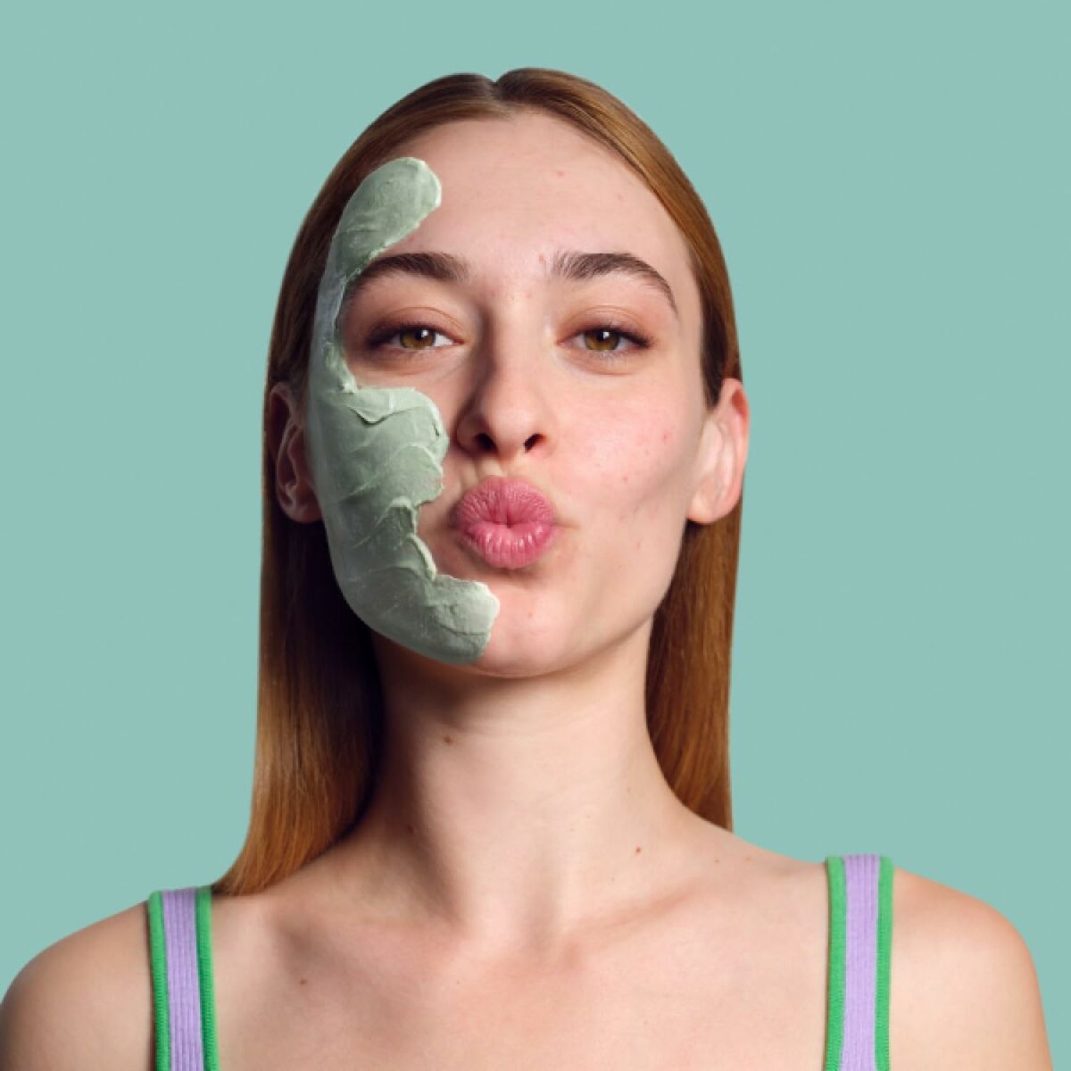 Young woman applying a green facial mask while making a playful kissy face, showcasing skincare routine.
