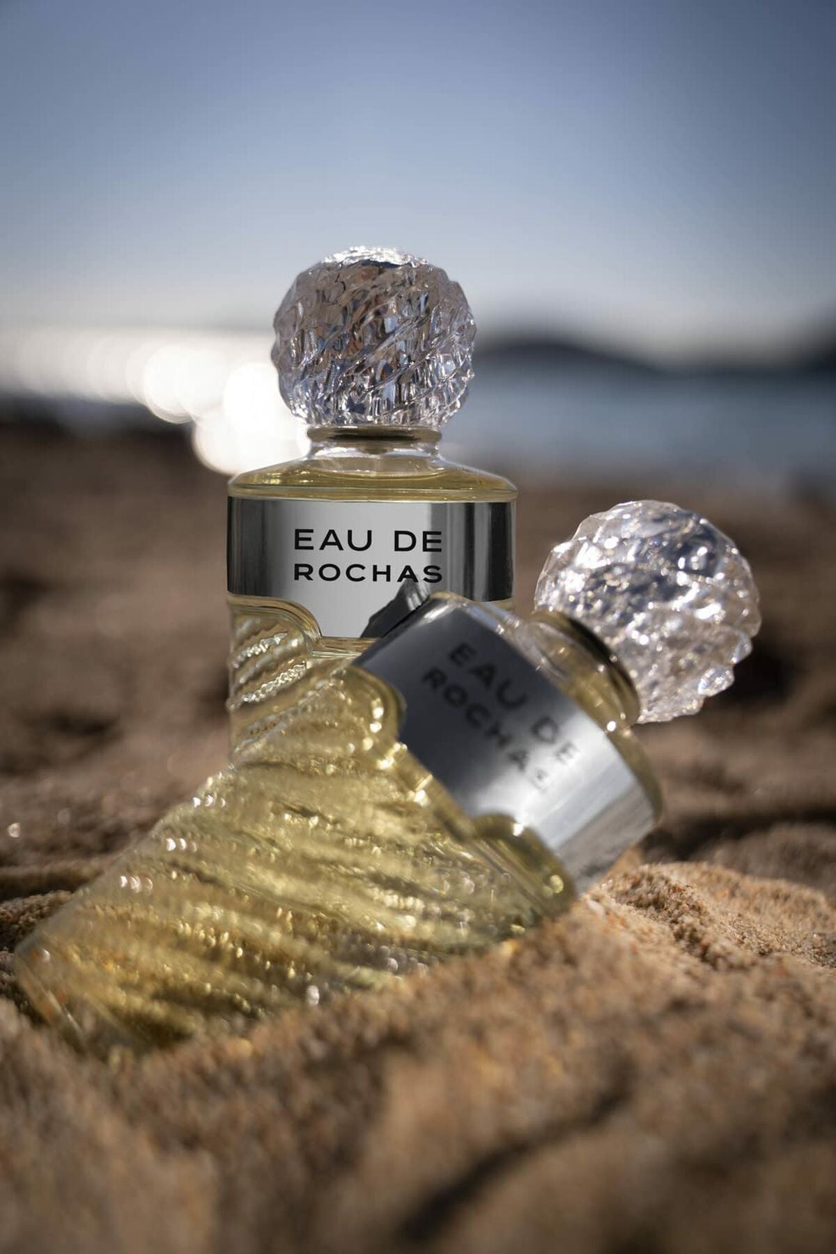 Bottles of Parfum Femme Rochas Eau de Rochas on sand, highlighting the elegant design and freshness of the fragrance.