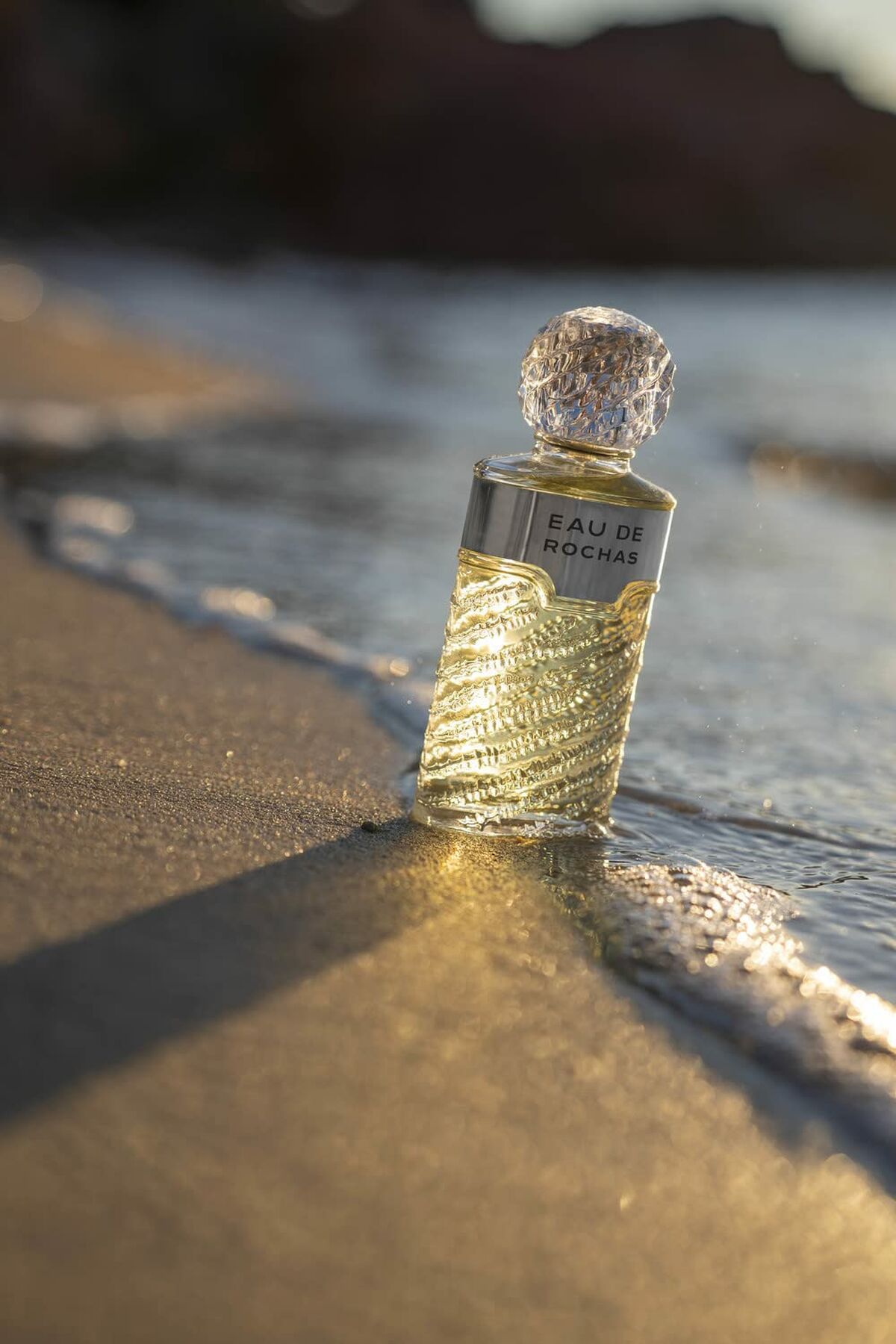 Parfum femme Rochas Eau de Rochas EDT 100ml posé sur le sable au bord de l'eau, lumière douce du coucher de soleil.