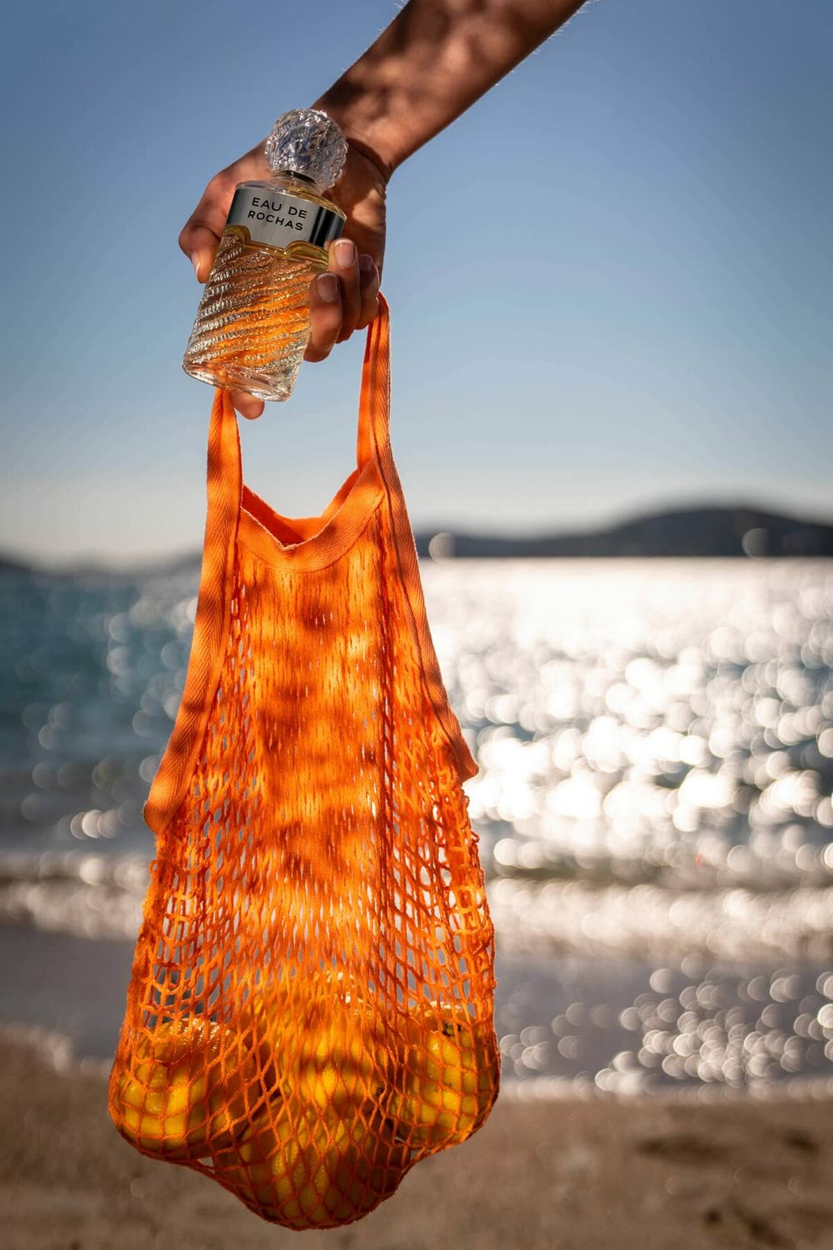 A hand holding an orange mesh bag with a bottle of Eau de Rochas against a sparkling beach background.
