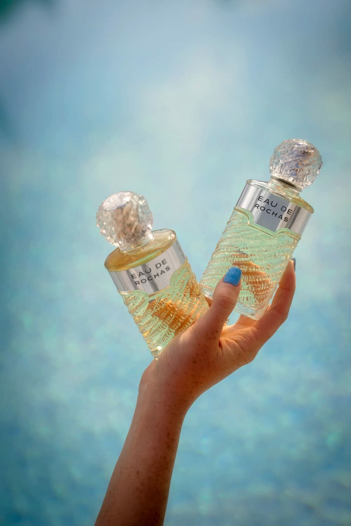 Parfum Femme Rochas RC001A01 EDT bottles held by hand against a shimmering blue background.