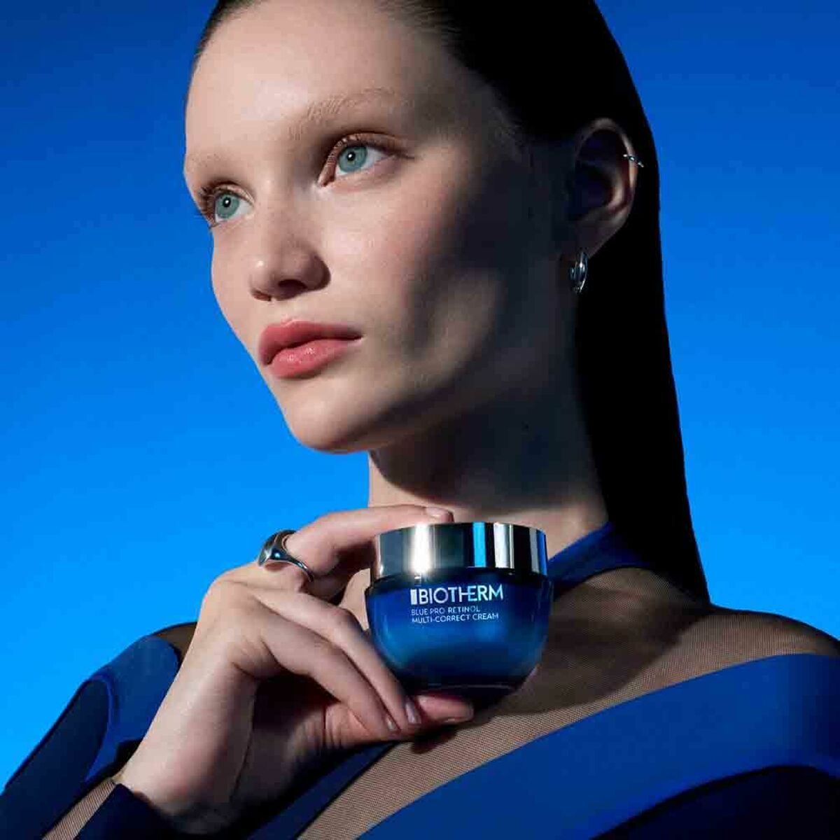 Model showcasing Biotherm Blue Retinol facial cream against a blue background, highlighting its luxurious texture and unisex appeal.