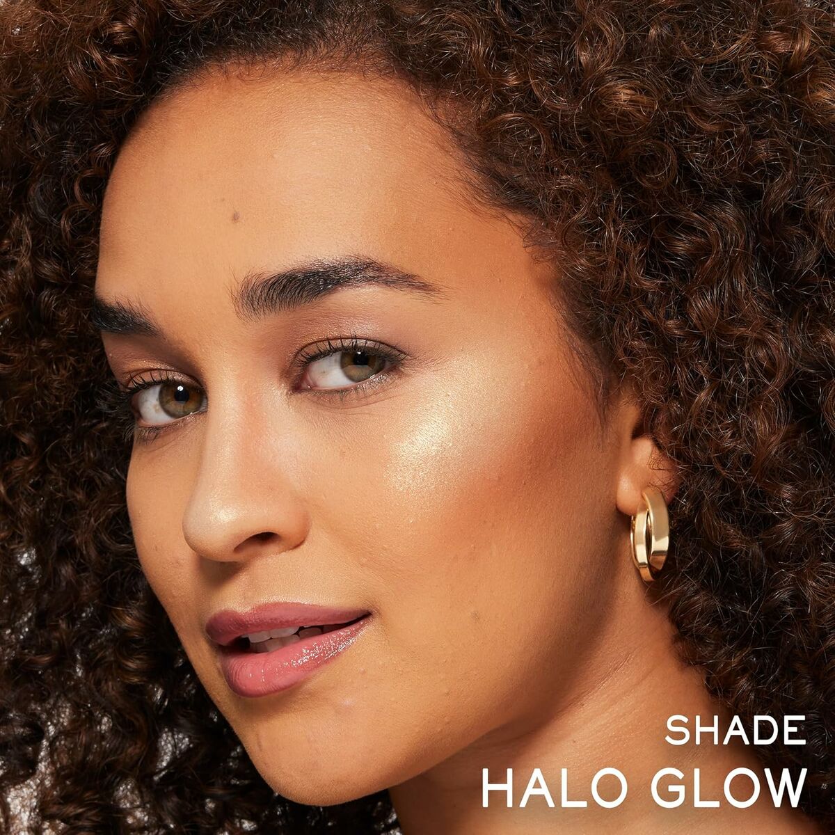Close-up of a model showcasing Halo Glow makeup shade with curly hair and gold hoop earrings.