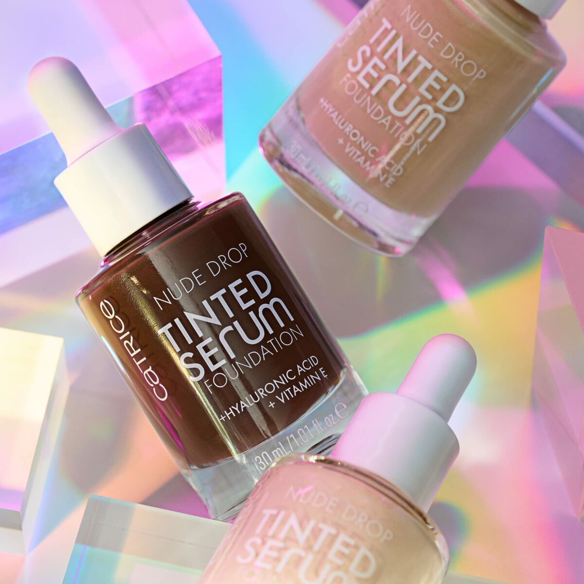 Catrice Nude Drop Tinted Serum Foundation bottles in various shades with hyaluronic acid and vitamin E, set against a colorful background.