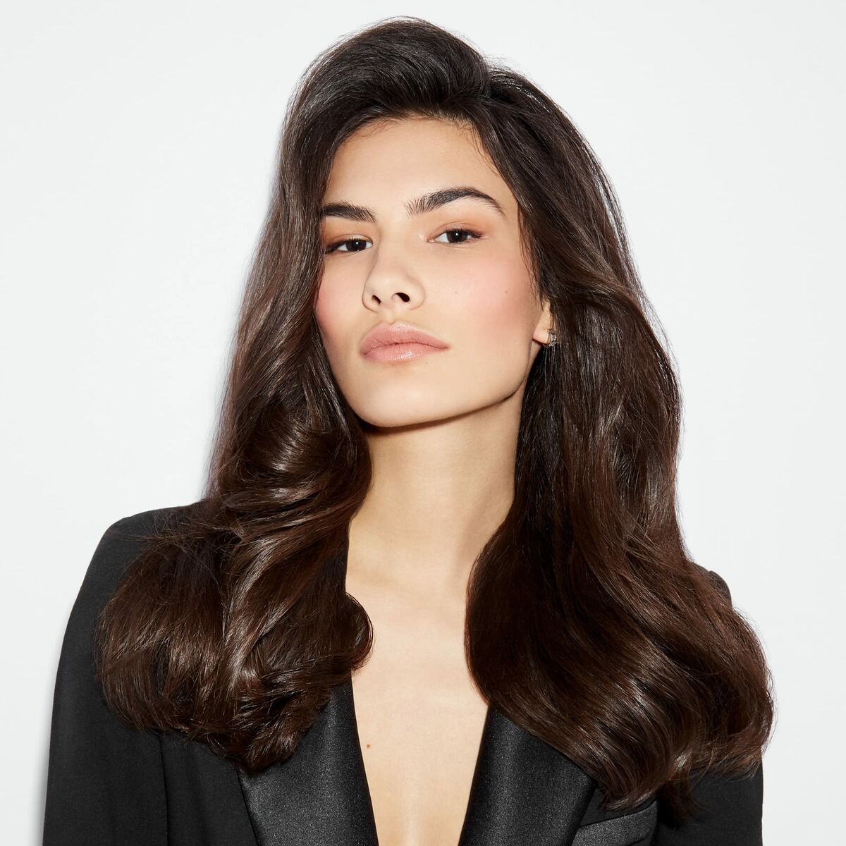 Femme aux cheveux brillants et volumineux portant un blazer noir sur fond blanc.