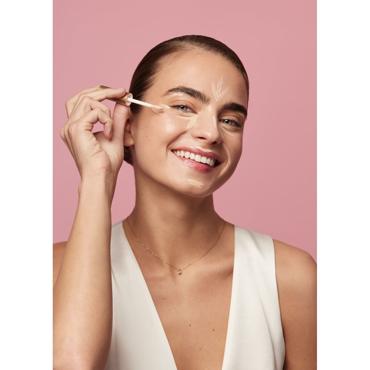 Woman applying Éclaircissant Sensilis SKIN GLOW FACIAL with a joyful smile against a pink background.