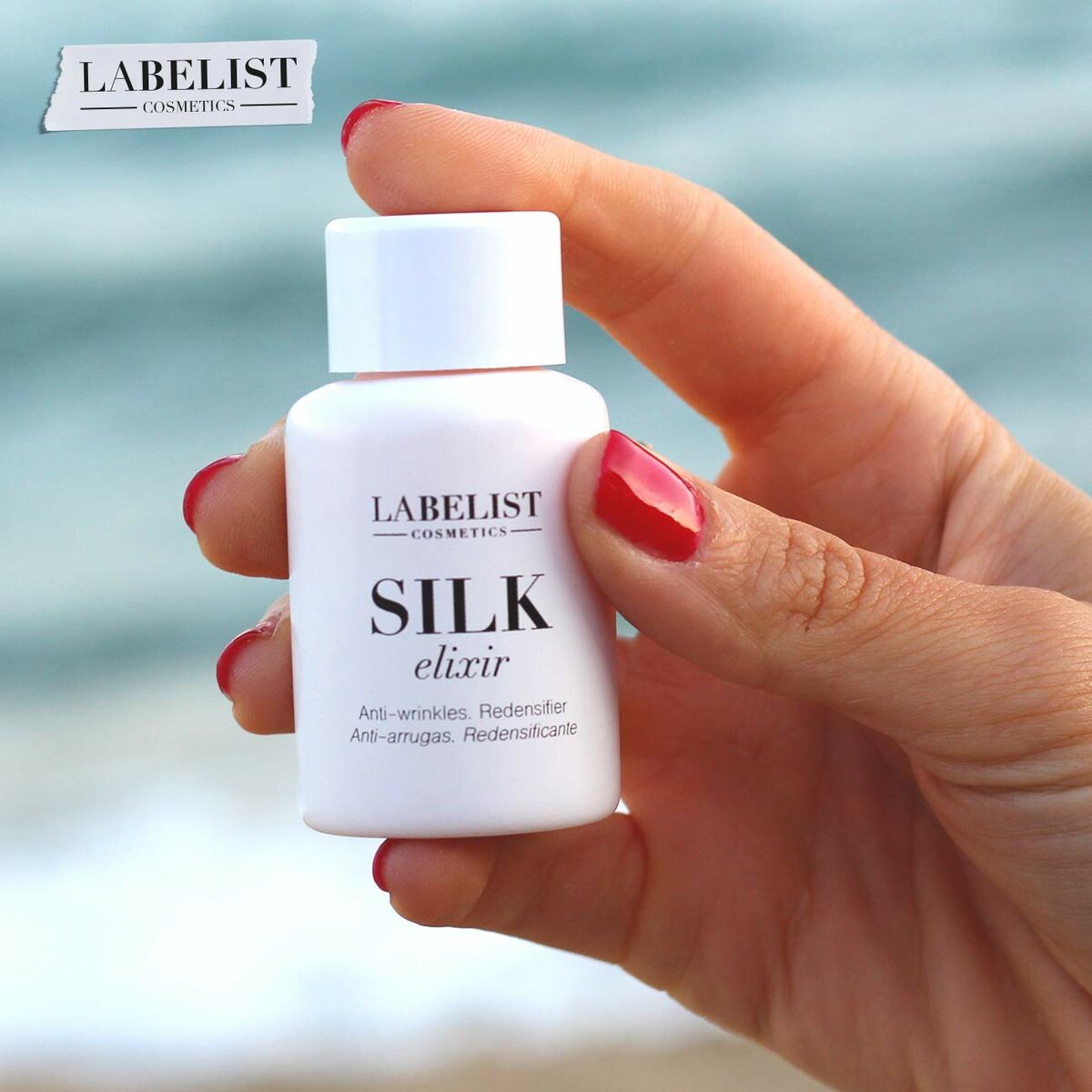 Bouteille de crème visage Labelist Cosmetics Silk Elixir, anti-rides, tenue en main, près de la plage.